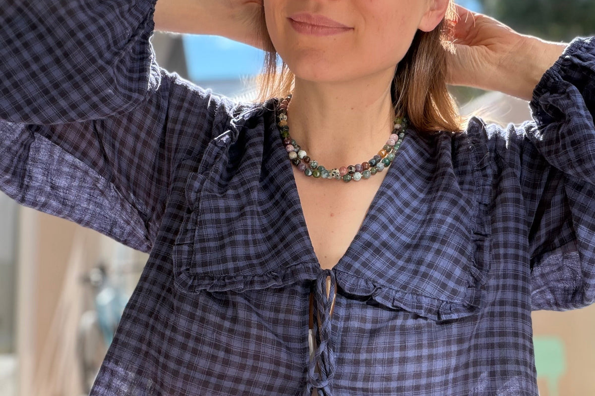 A woman wearing a dark purple and black plaid long-sleeve blouse with a ruffled collar and front tie, paired with white pants, photographed outdoors on a sunny day.