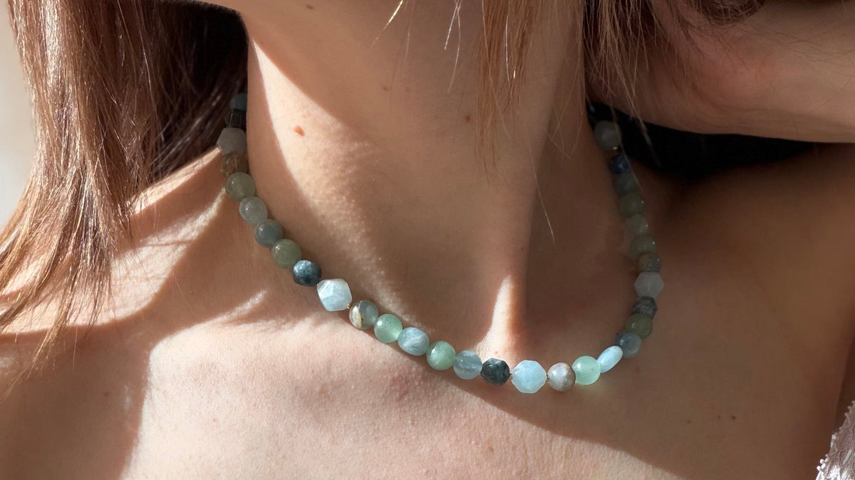 A close-up of a woman wearing a beaded necklace made of light blue, mint green, and gray round beads, paired with a light beige knit top.