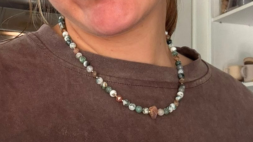 A close-up selfie of a woman wearing a brown crewneck sweatshirt and a beaded necklace in a kitchen setting.
